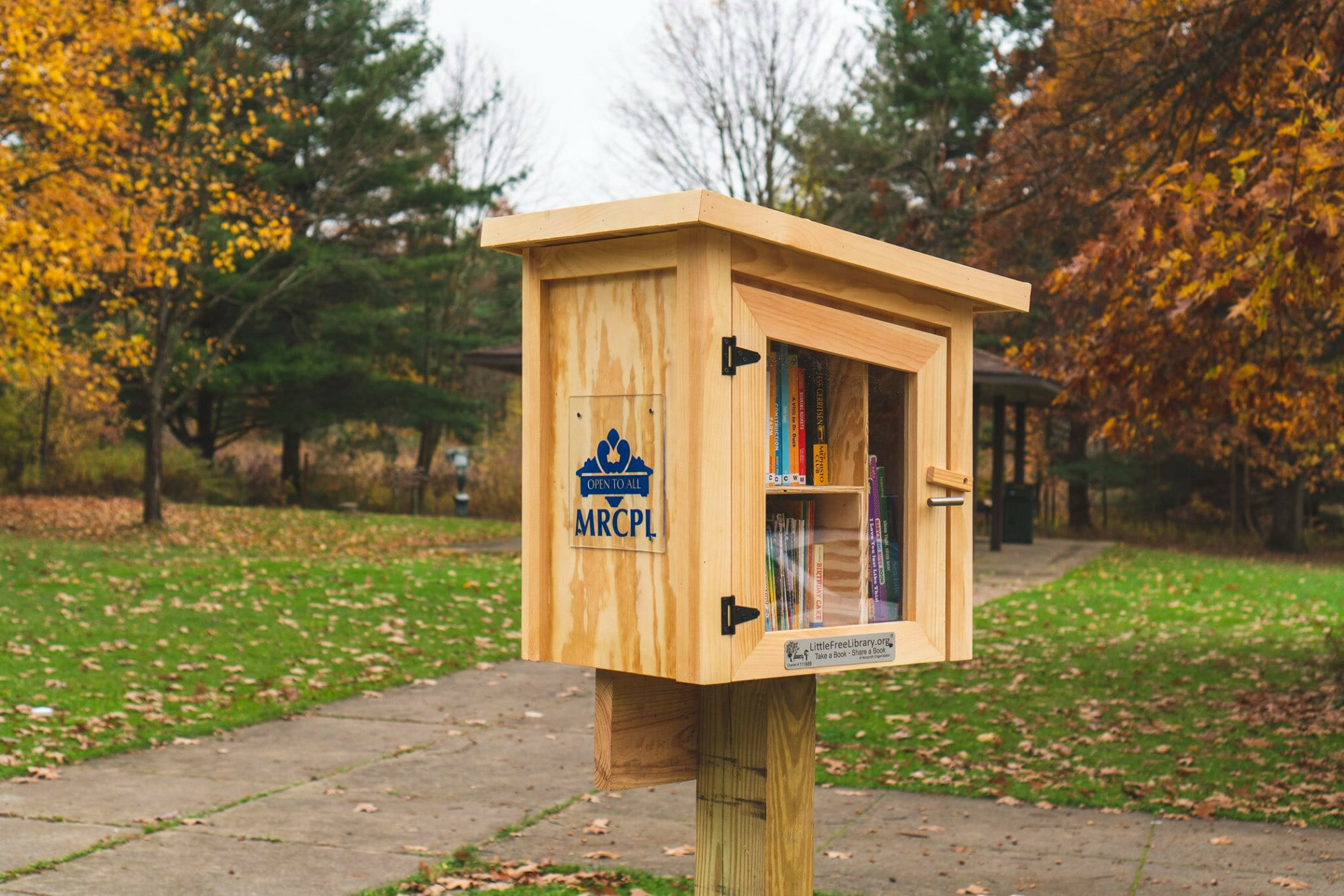 a wooden, MRCPL little free library box stands outdoors in the fall at a park.