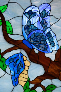 Stained glass window featuring a blue butterfly on a branch of a tree with a blue and cream chrysalis hanging on the branch.