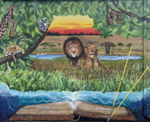 An open book with a jungle scene coming out of it. There are lions, giraffes, cheetahs, koalas, elephants, monkeys, and zebras enjoying the nature at sunset. Painted by Robin Shoup Wilson.