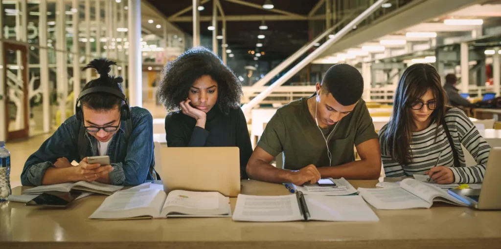 Students studying in a library with books and a laptop.