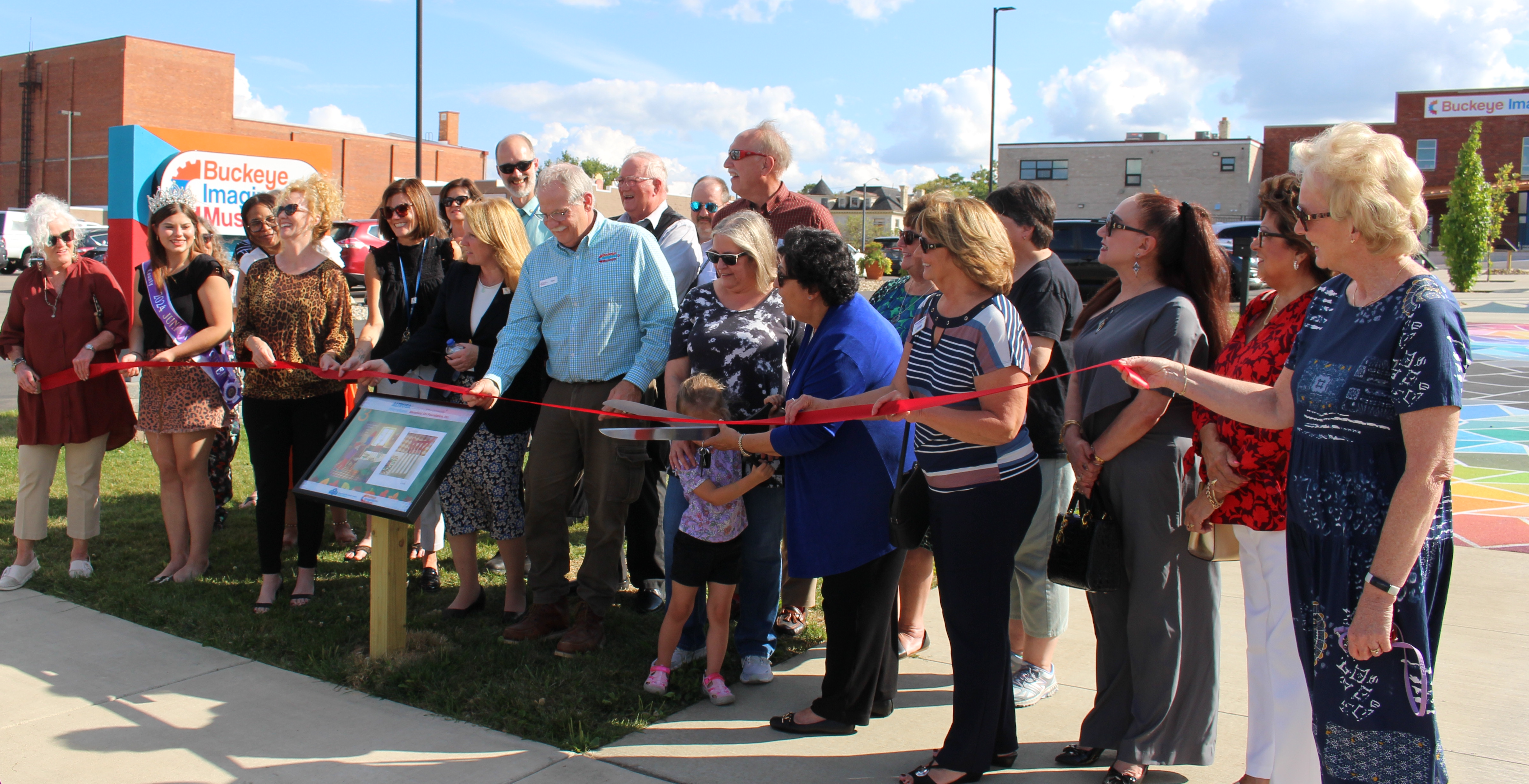 Group of people stand outdoors at a StoryWalk station at a ribbon cutting ceremony.